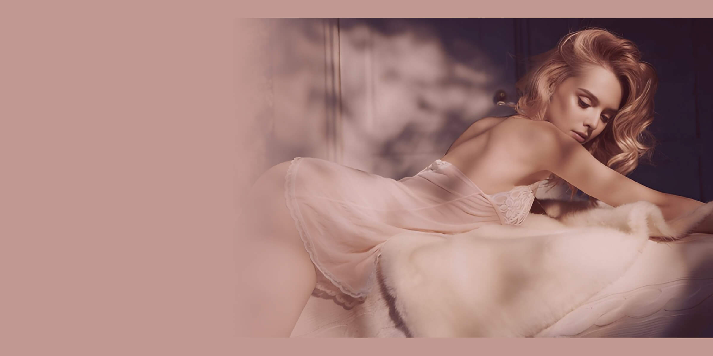 Blonde model reclining in a sheer blush-pink lace-trimmed chemise, draped over soft fur in a warm, dreamy boudoir setting.