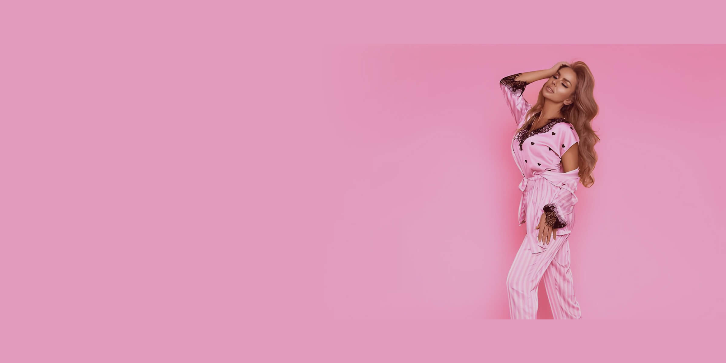 Blonde model in a pink striped satin pyjama set with black lace trim, posing against a soft pink backdrop.